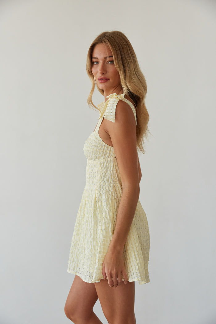 A woman wearing a white and yellow gingham mini dress with bow tie straps and a flared silhouette.