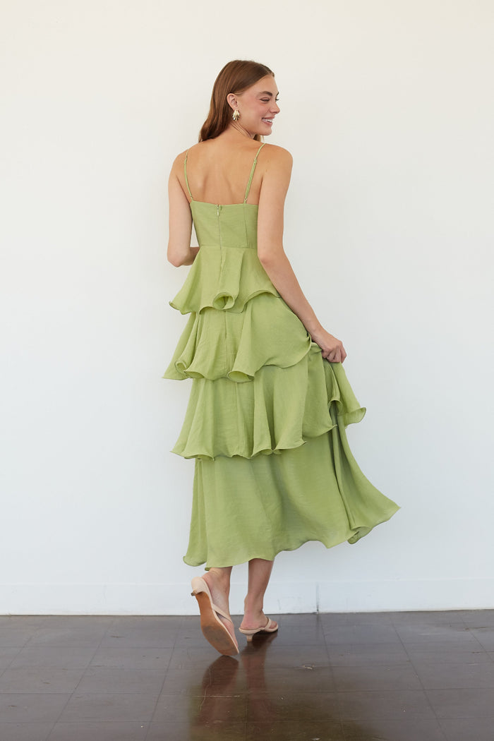 A woman wearing a light green tiered maxi dress, shown from the back as she walks against a plain white wall.