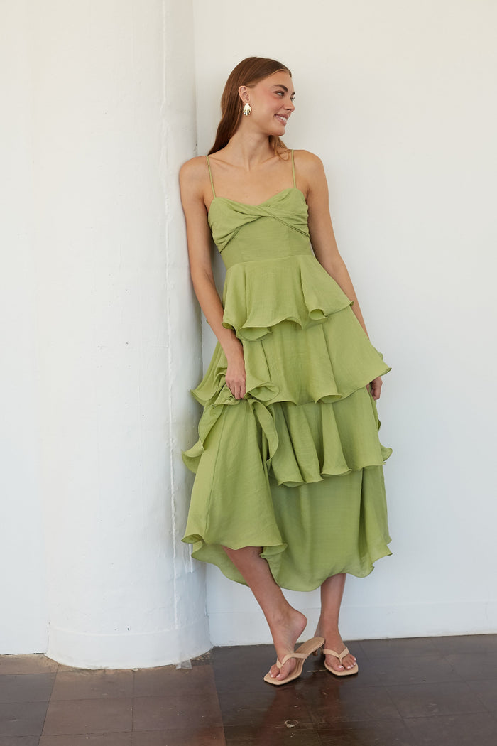A woman wearing a light olive green strapless ruffled midi dress with a twisted sweetheart bodice, leaning against a white wall.