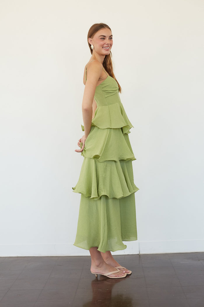 A woman modeling a light green tiered maxi dress with side cutouts, standing in profile against a plain white wall.