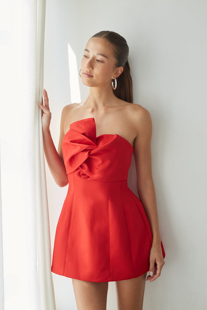 red-image | front | Woman wearing a red strapless dress with a large bow detail, standing against a white wall.