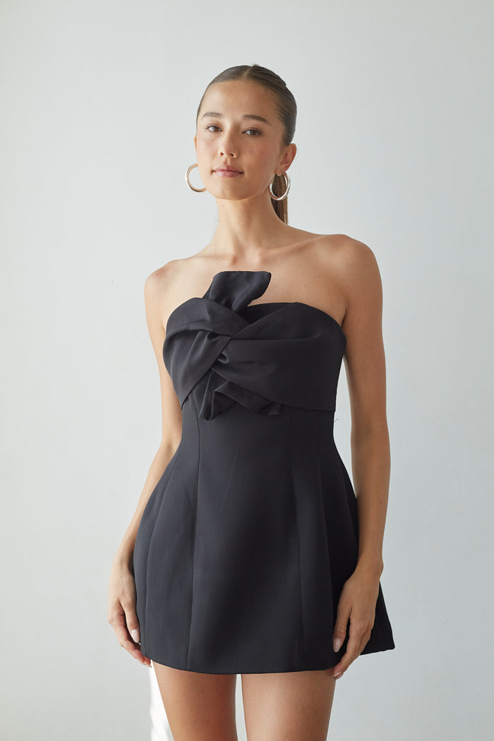Woman wearing a black strapless dress with a knot detail on a plain background