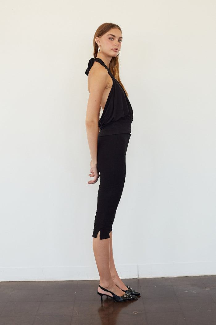 A woman stands in profile wearing a black sleeveless halter-neck top and matching black capri-length skirt or dress, paired with black pointed flats, against a plain white wall and dark floor.