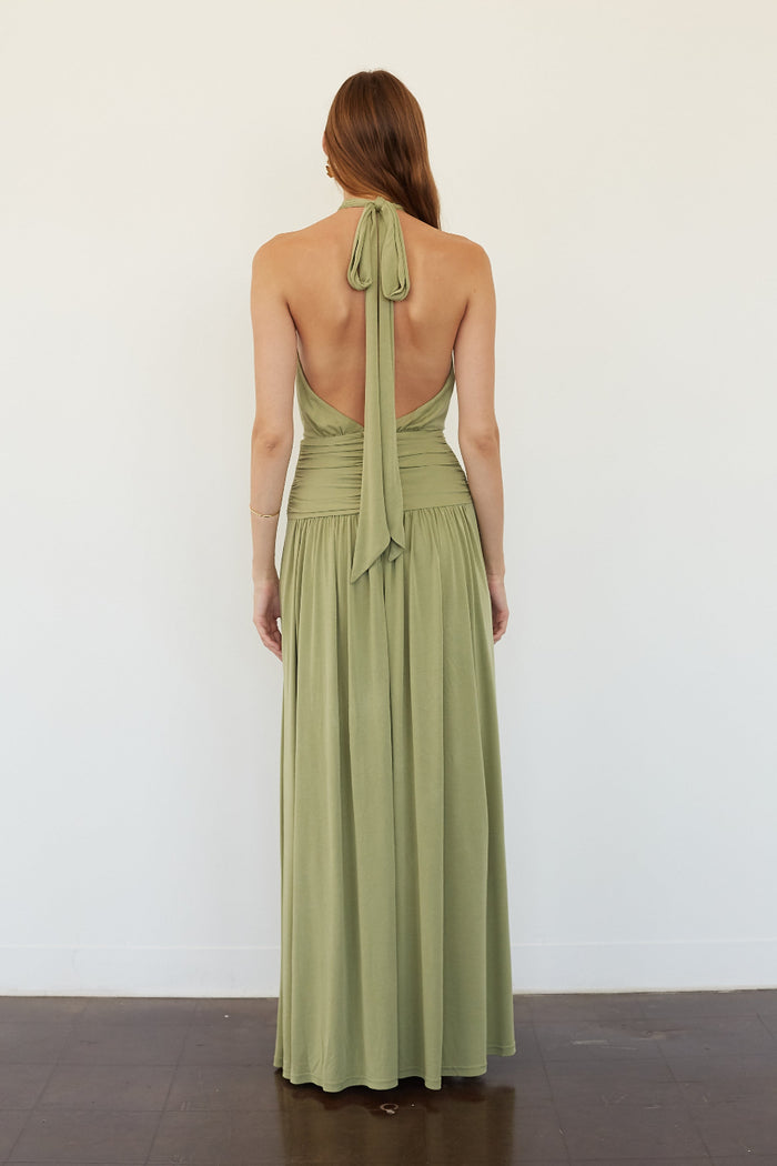 A woman stands with her back to the camera wearing a sage green halter-neck maxi dress with a tie-back detail and an open back, set against a plain white wall.