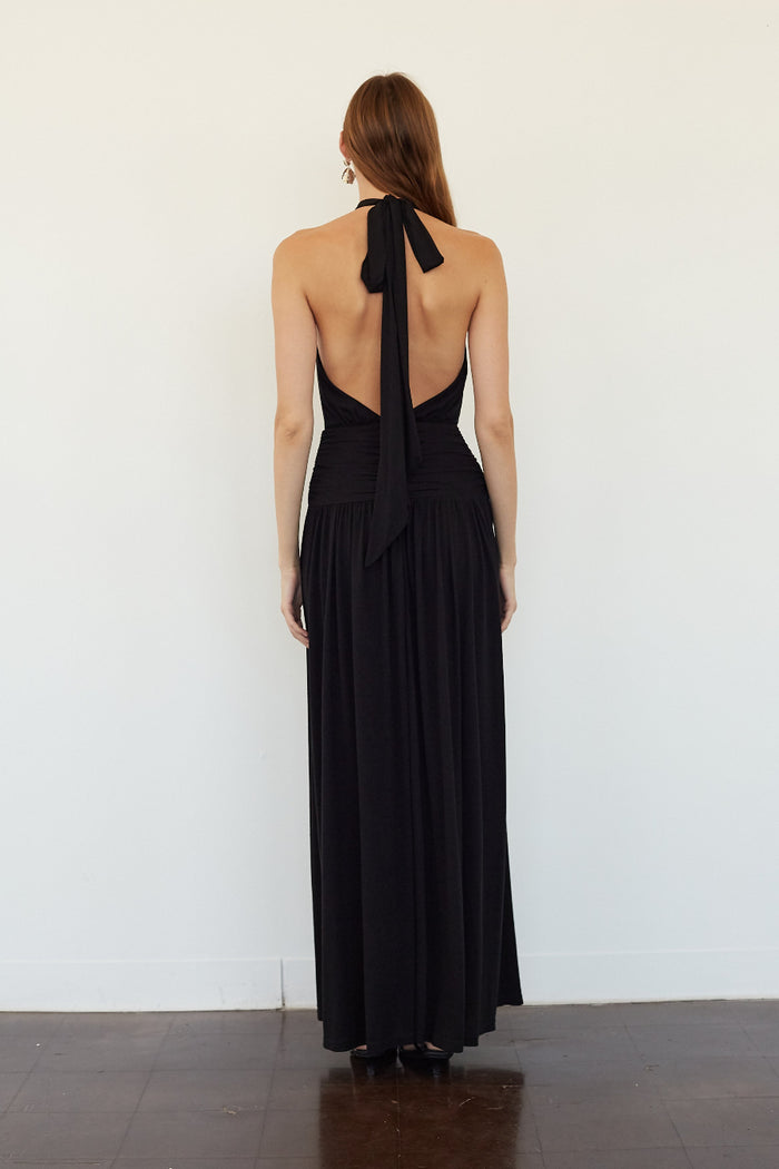 A woman viewed from the back wearing a black halter-neck maxi dress with a deep open back and long tie detail, standing against a plain white wall.