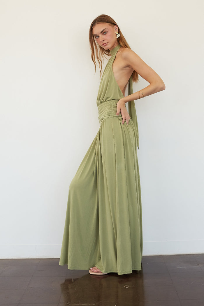 A woman modeling a sage green halter-neck maxi dress with a wrap-style waist and flowing skirt, standing against a plain white wall.