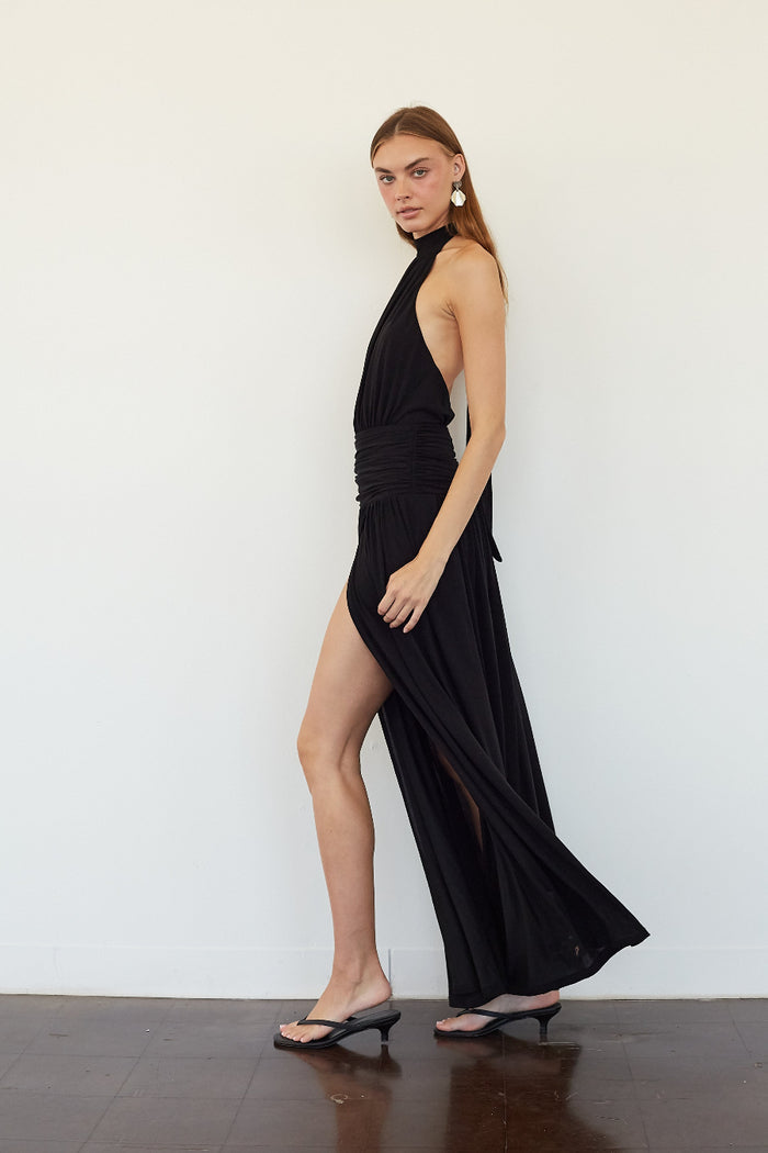 A woman models a black halter-neck maxi dress with a high thigh slit and draped side detail, paired with black thong sandals, standing against a plain white wall.