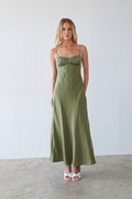 green-image | A woman modeling a long olive green satin maxi dress with thin spaghetti straps and a ruched sweetheart neckline, standing against a neutral light gray wall and concrete floor.
