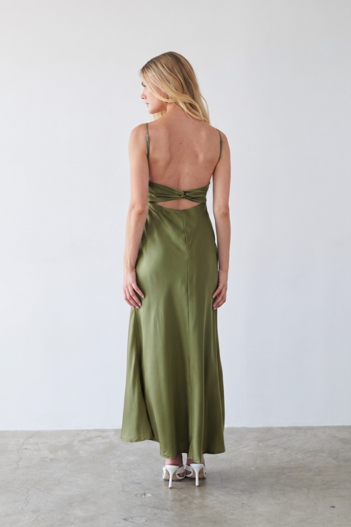 A woman stands with her back to the camera wearing a satin-like olive green maxi dress with thin straps and a cutout at the back, paired with white high heels, in a minimalist studio setting.