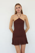 A woman modeling a dark brown halter-neck mini dress with a ruched bodice and flared ruffled hem, set against a plain white background.