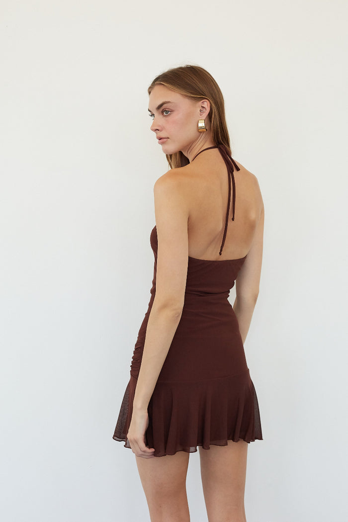 A woman modeling a dark brown halter mini dress from the back, featuring a ruched bodice and a flared ruffled hem, set against a plain white background.