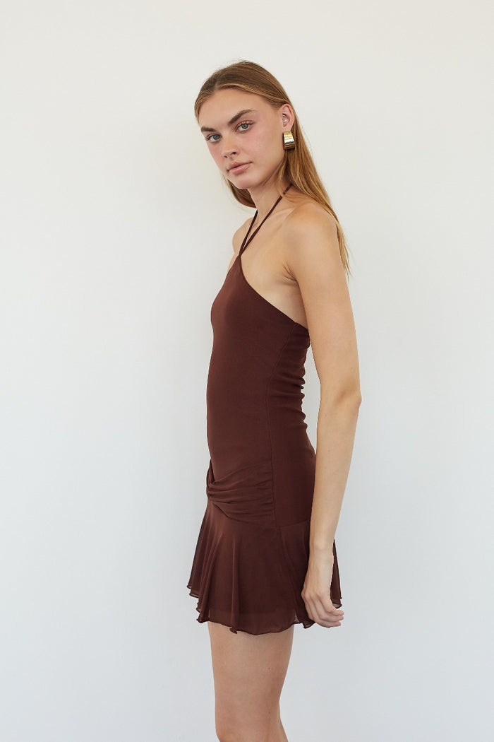 A woman modeling a dark brown halter-neck mini dress with a ruched bodice and flared ruffled hem, set against a plain light gray studio background.