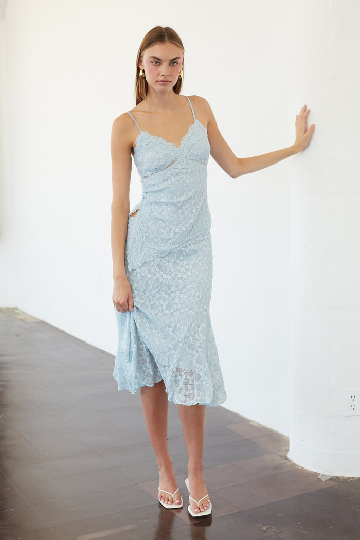 A woman modeling a light blue lace spaghetti-strap midi dress in a minimalist white-walled room with dark wood floors.