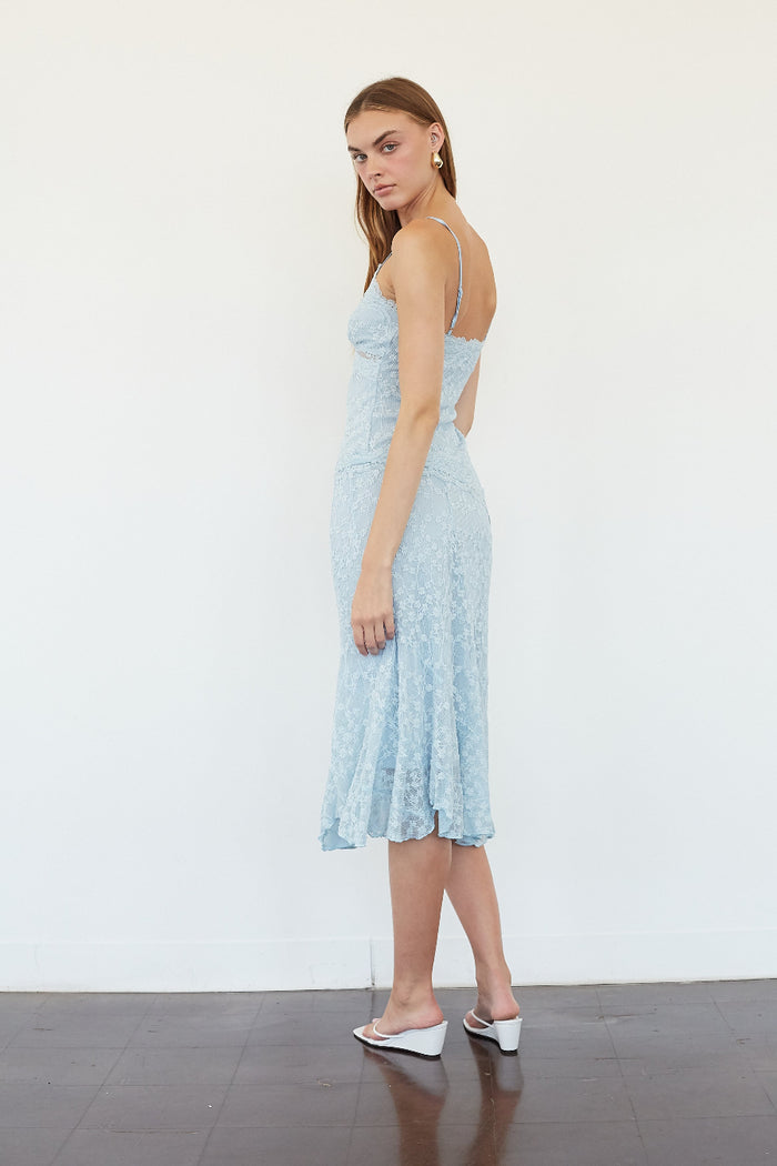 A woman modeling a light blue lace midi dress with spaghetti straps and a back cutout, standing against a plain white wall.