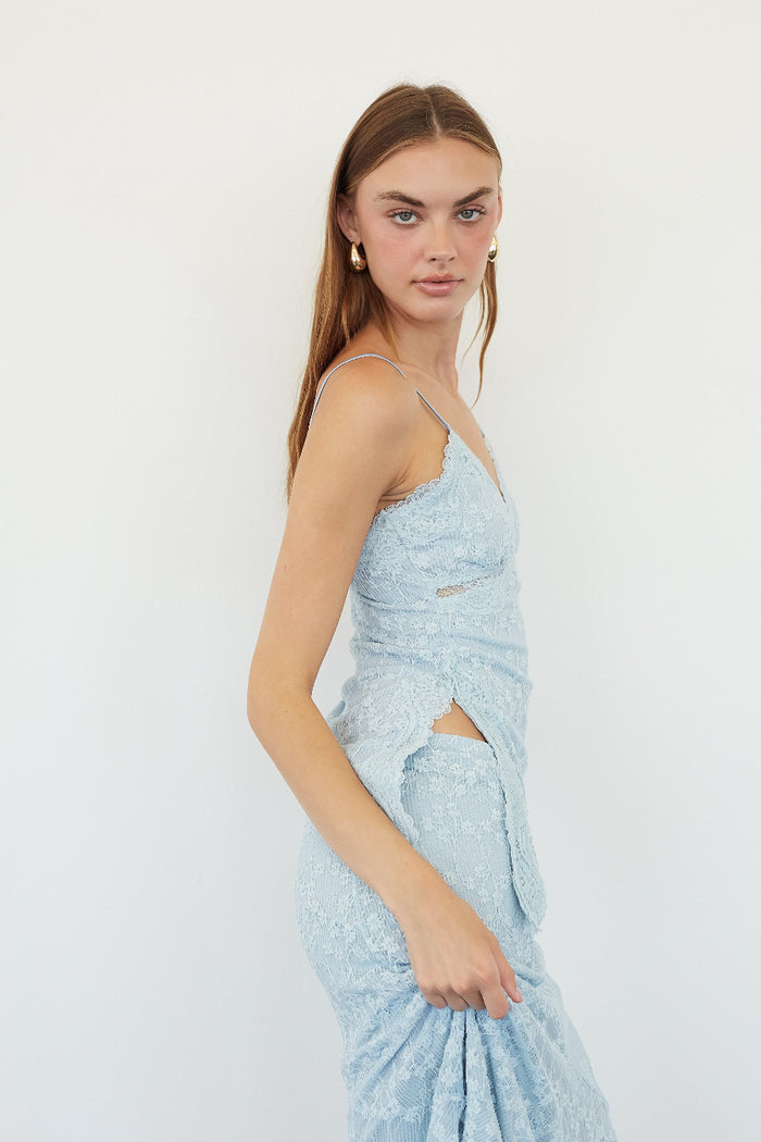 A woman modeling a light blue lace camisole dress with spaghetti straps, side cutouts, and a matching skirt, photographed against a plain white studio background.