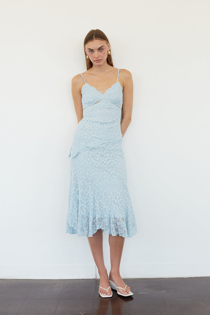 A woman modeling a light blue lace midi dress with thin straps, a sweetheart neckline, and a ruffled hem, standing against a plain white wall.