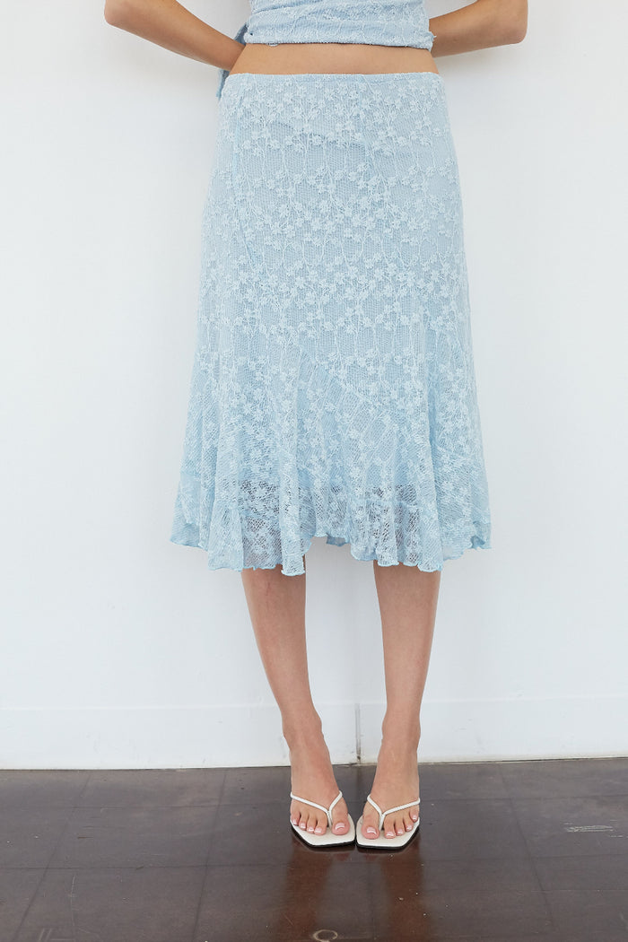A light blue floral lace midi skirt worn by a model, paired with a matching light blue crop top and white strappy heels, standing against a white wall.
