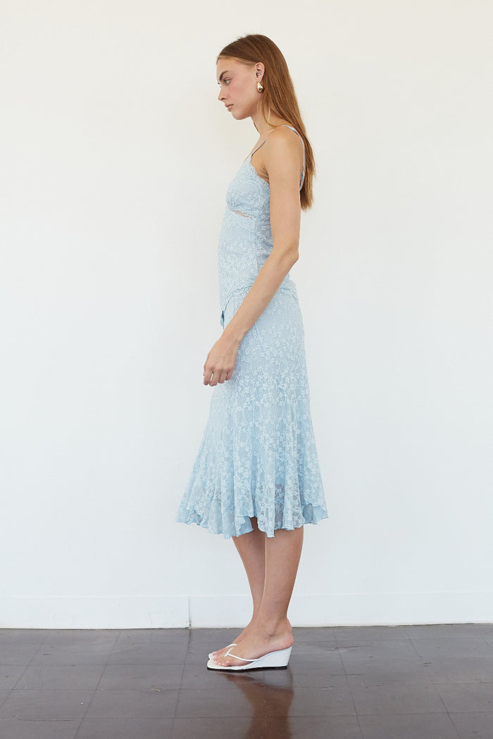 A woman stands in profile wearing a light blue sleeveless lace midi dress with a flared hem, paired with white wedge sandals, against a plain white wall and dark floor.