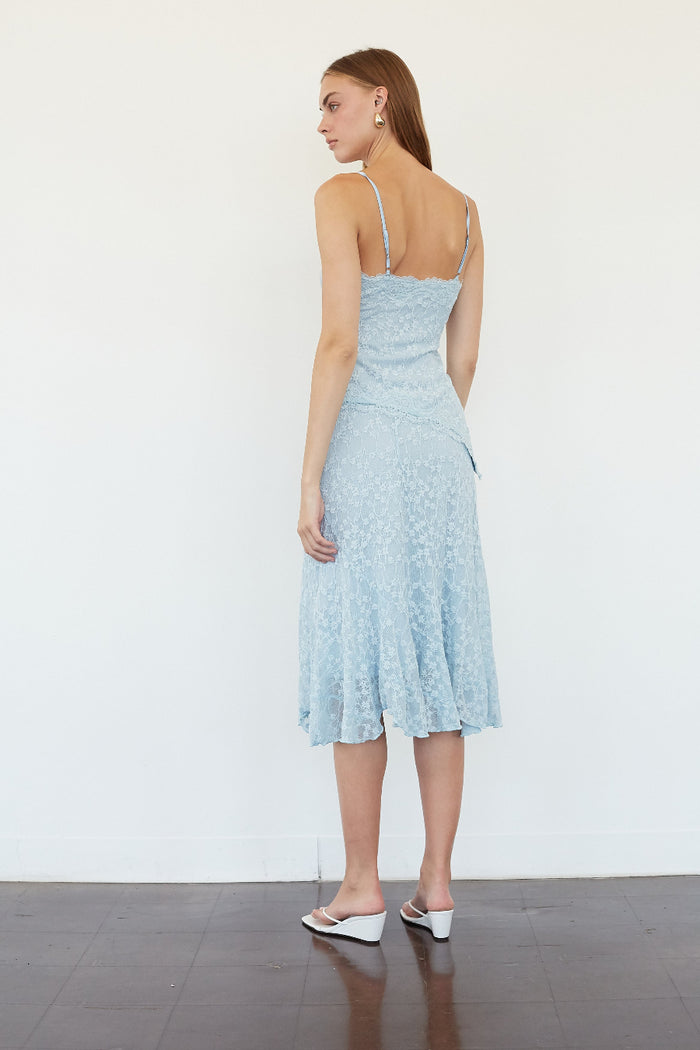 A woman stands with her back to the camera wearing a light blue lace midi dress with spaghetti straps and white wedge sandals against a plain white wall.