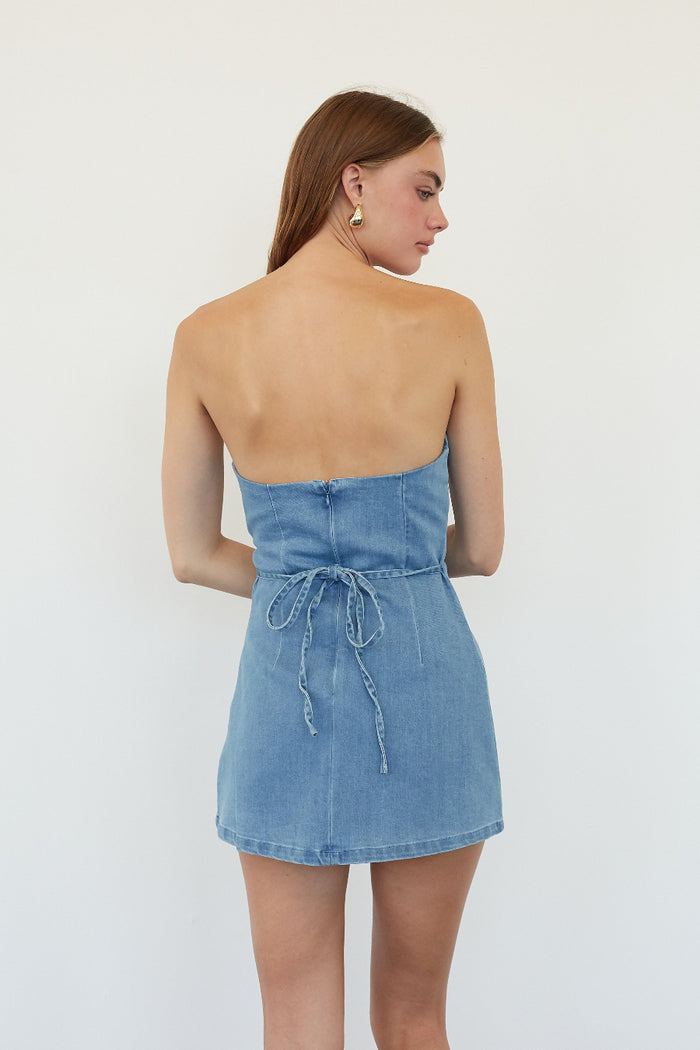 A woman shown from the back wearing a light blue denim strapless mini dress with a tie detail at the waist, set against a plain white studio background.