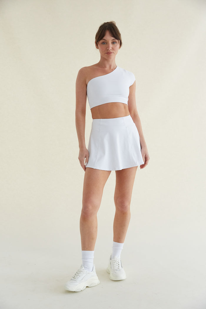 front view cream seamless background with a model wearing a one shoulder cropped athleisure top and a matching flared mini skort with pockets - crew socks and chunky dad sneakers - women's athleisure