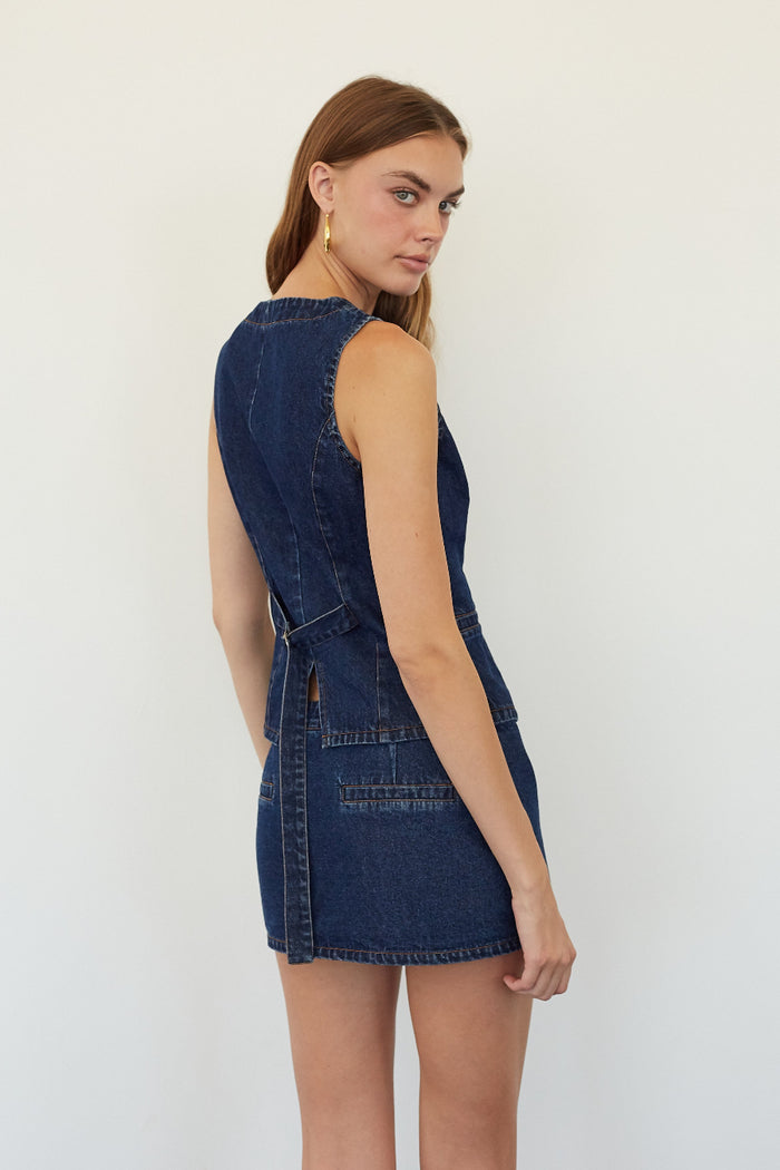 A woman modeling a dark blue denim sleeveless mini dress from the back, standing against a plain white studio background.