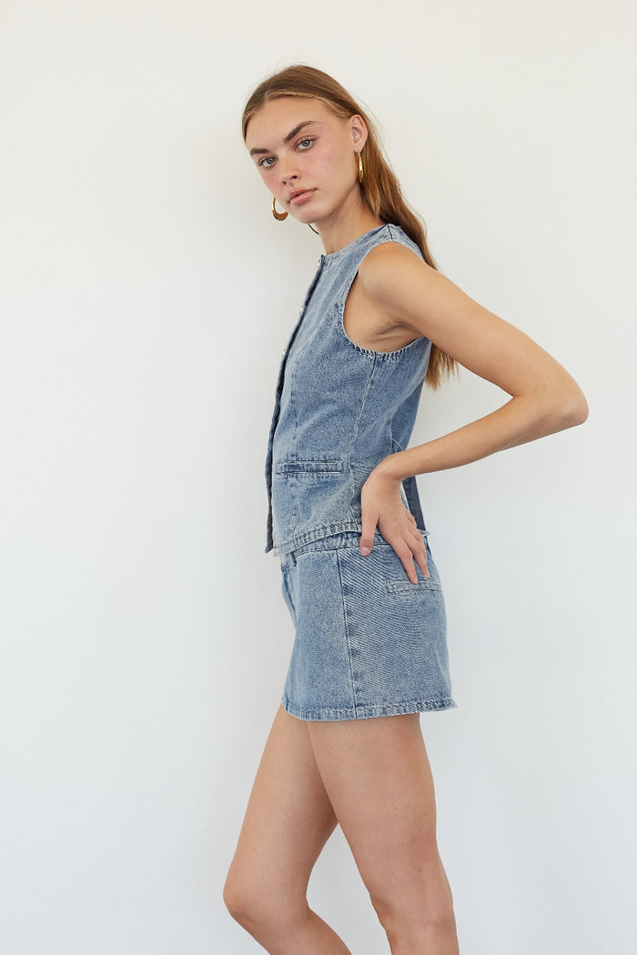 A woman modeling a light blue denim overall-style romper or short dress with a sleeveless front, standing against a plain white studio background.