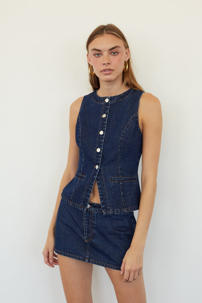 A model wearing a dark blue denim two-piece set consisting of a sleeveless button-front top and a matching mini skirt, set against a plain white background.