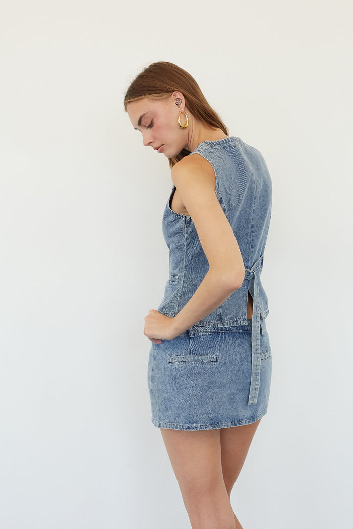 A woman wearing a sleeveless light blue denim mini dress, shown from a side-back angle against a plain white studio background.