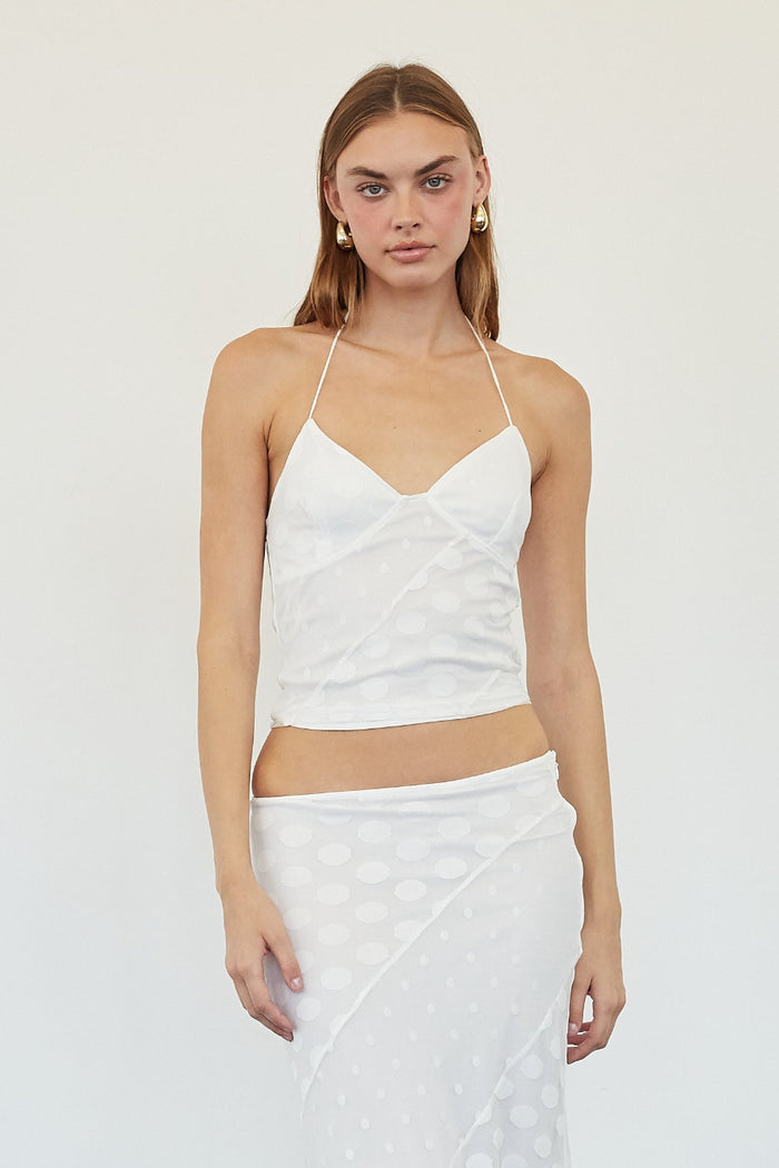 A woman modeling a white two-piece outfit consisting of a sleeveless crop top with thin straps and a matching long skirt with a polka dot pattern, standing against a plain white wall.