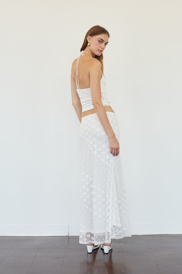 A woman modeling a white two-piece outfit consisting of a halter crop top and a matching long sheer skirt with a polka dot pattern, standing against a plain white wall.