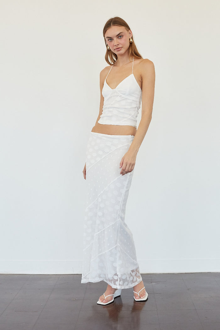 A woman modeling a white two-piece outfit consisting of a textured camisole top and a matching sheer patterned maxi skirt, standing against a plain white wall.