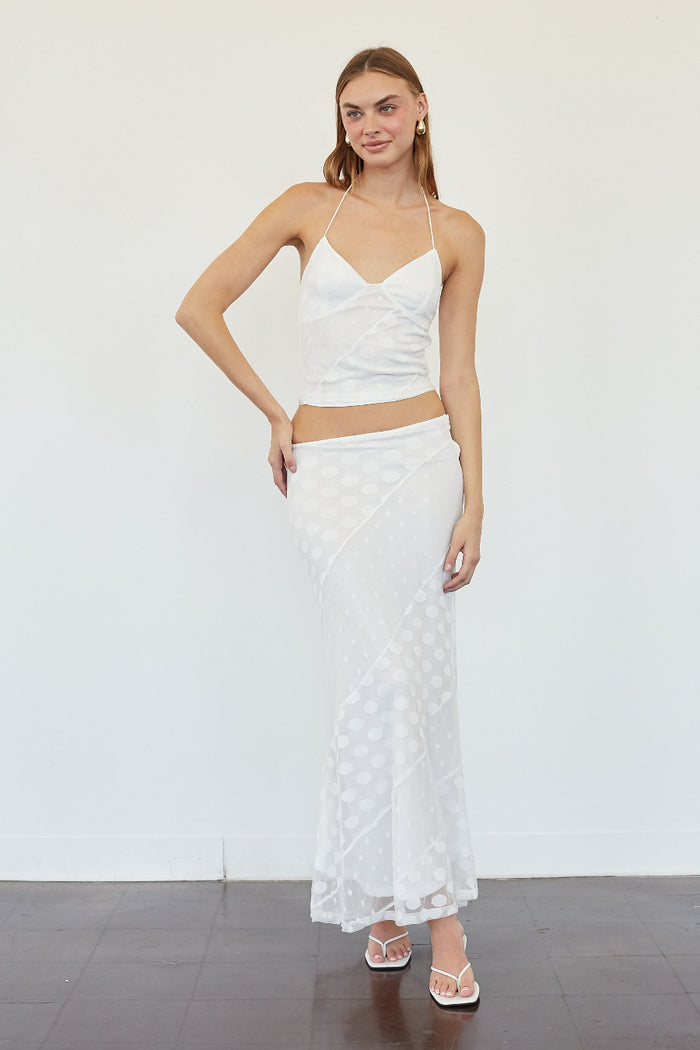 A woman modeling a white two-piece outfit consisting of a spaghetti-strap crop top and a matching textured midi skirt, standing against a plain white wall.