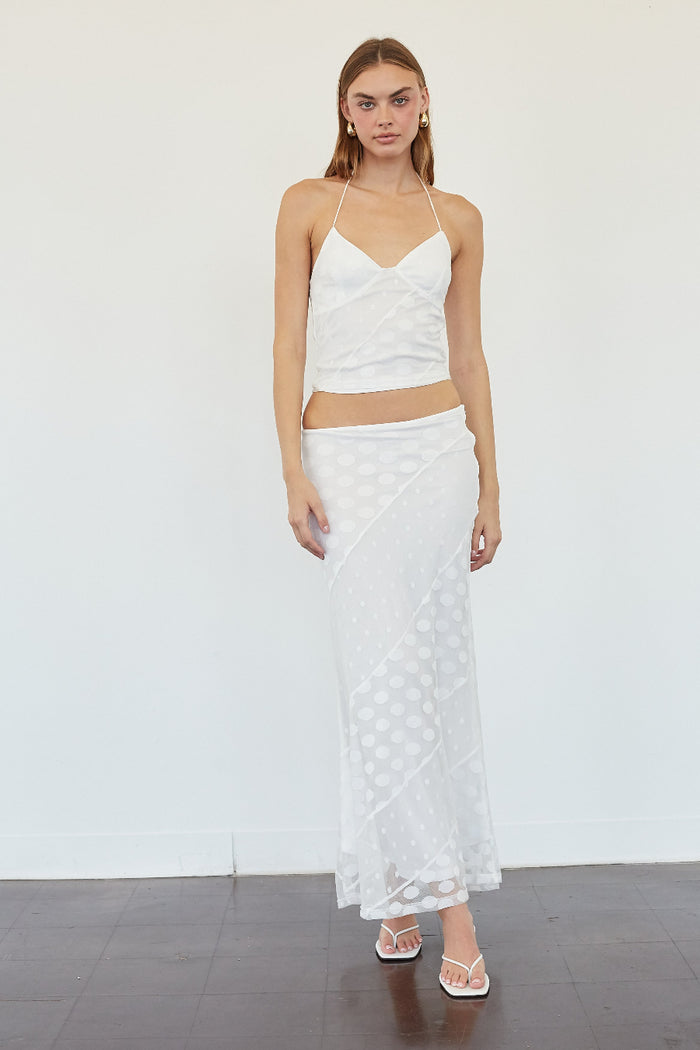 A woman modeling a white two-piece outfit consisting of a sleeveless crop top with thin straps and a matching long skirt with a polka dot pattern, standing against a plain white wall.