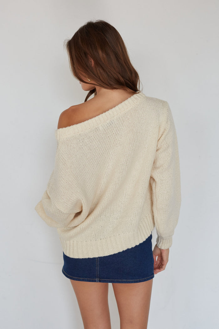 Woman wearing a beige off-shoulder sweater and blue skirt against a white background