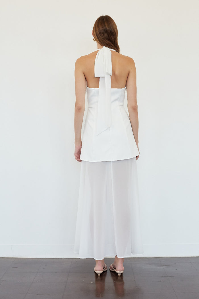 A woman viewed from the back wearing a white sleeveless halter dress with a large bow detail at the back and a flowing midi skirt, standing against a plain white wall.