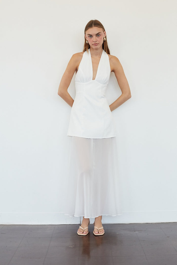 A woman modeling a white sleeveless halter dress with a deep V-neckline and a flowing sheer maxi skirt, standing against a plain white wall.