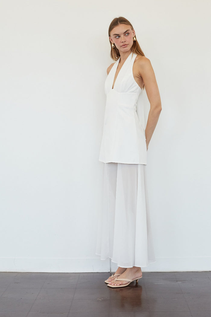 A woman modeling a white sleeveless halter-style top with a draped, layered skirt or wide-leg overlay, standing against a plain white wall on a dark wood floor.