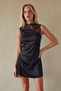 black-image | A woman modeling a black satin mini dress with a sheer black lace mock neck and lace trim at the hem, posed against a neutral beige textured wall.