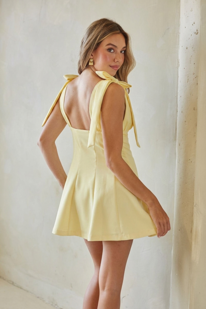 A woman modeling a pale yellow sleeveless mini dress with tie-shoulder straps and a flared skirt, photographed indoors against a textured off-white wall.