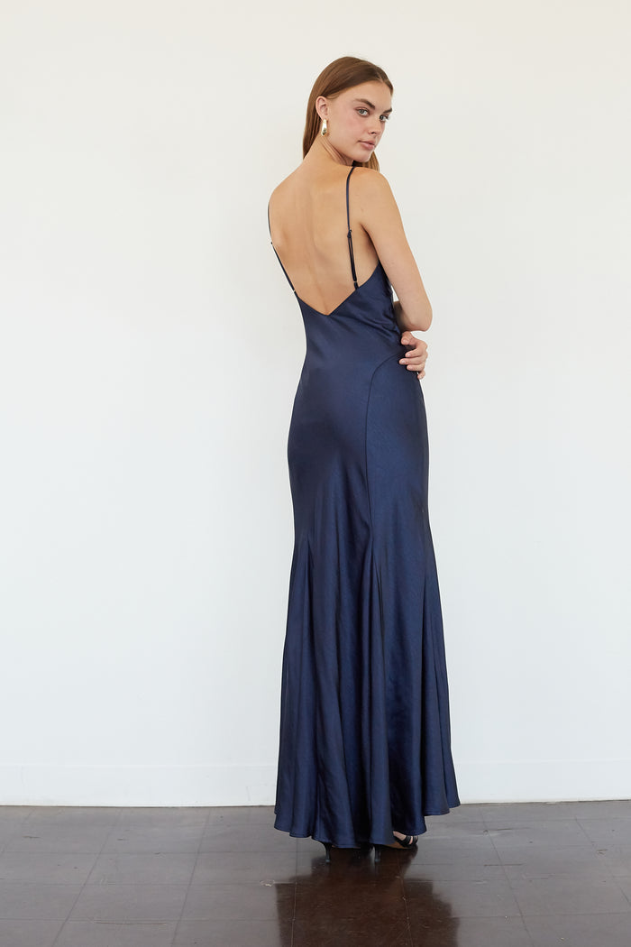A woman modeling a navy blue satin maxi dress from the back, featuring a low open back and thin straps, set against a plain white wall.
