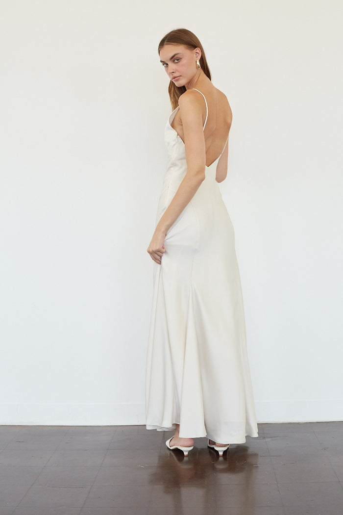 A woman modeling a floor-length white satin gown from the back, showing a low open back and thin straps, standing on a dark wood floor against a white wall.