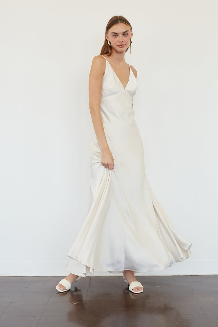 A woman modeling a sleeveless, off-white satin maxi dress with a deep V-neckline and flowing skirt, standing against a plain white wall.