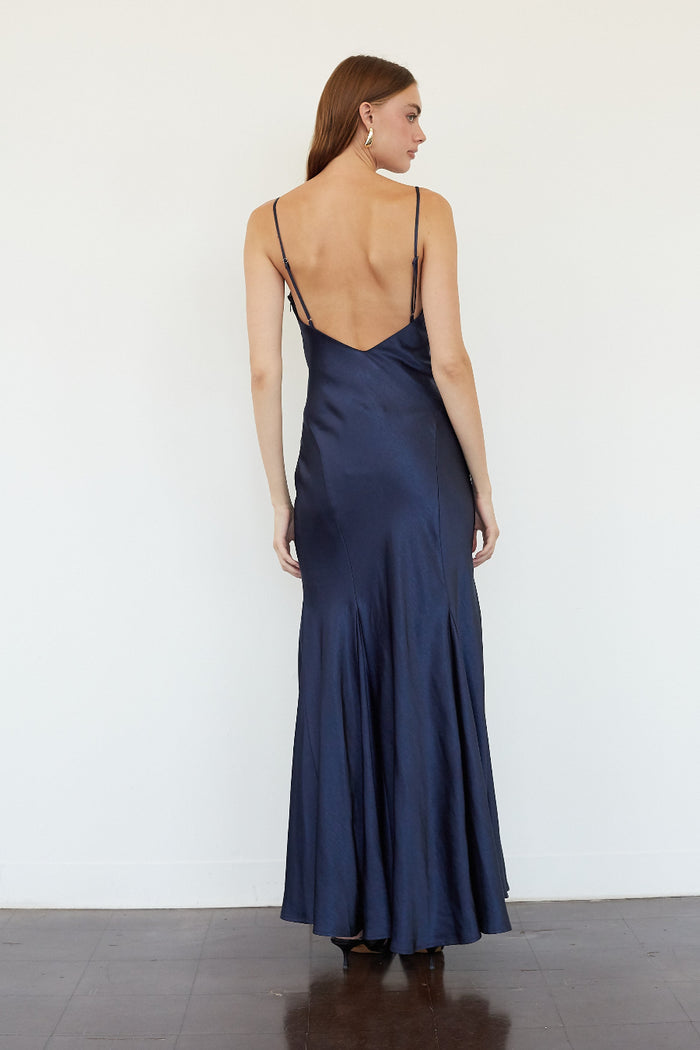 A woman stands with her back to the camera wearing a floor-length navy blue satin slip dress with thin spaghetti straps and a low open back, set against a plain white wall.