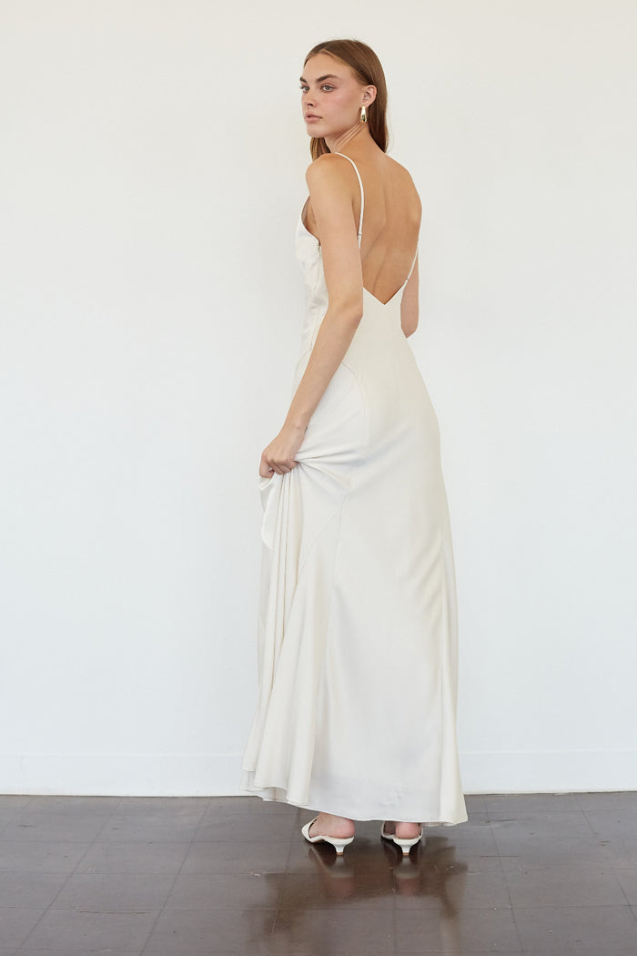 A woman modeling a floor-length white satin slip dress with thin straps and an open back, shown from the back in a minimalist studio setting.
