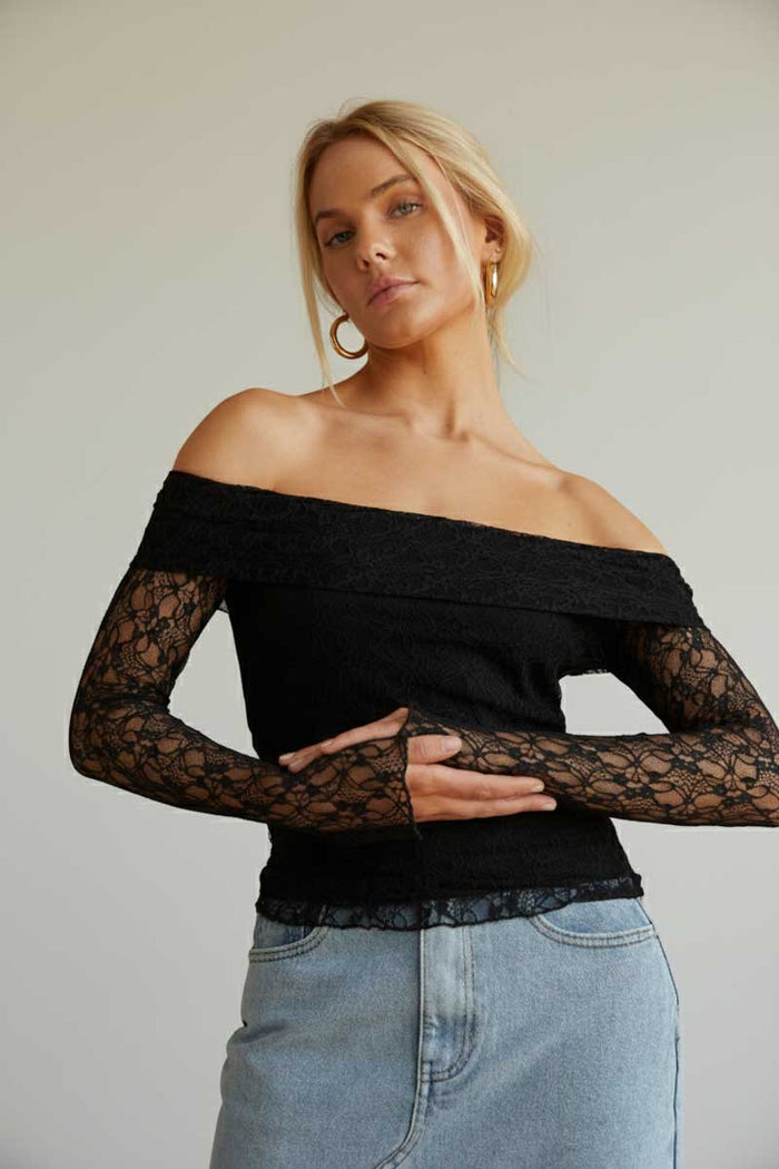 Woman wearing a black off-shoulder top with lace sleeves and light blue jeans against a plain background
