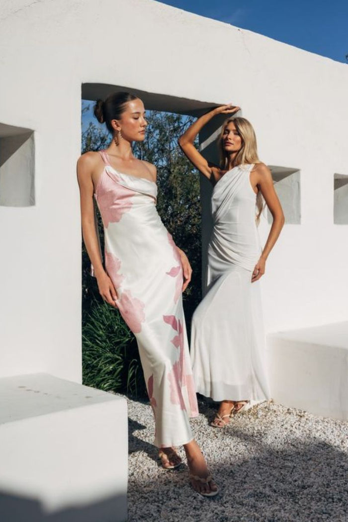 Two women in elegant dresses standing in a modern architectural setting with white walls and blue sky.