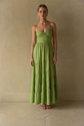 green-image | A woman stands barefoot against a textured beige wall wearing a sleeveless, tiered maxi dress in a light olive green color.