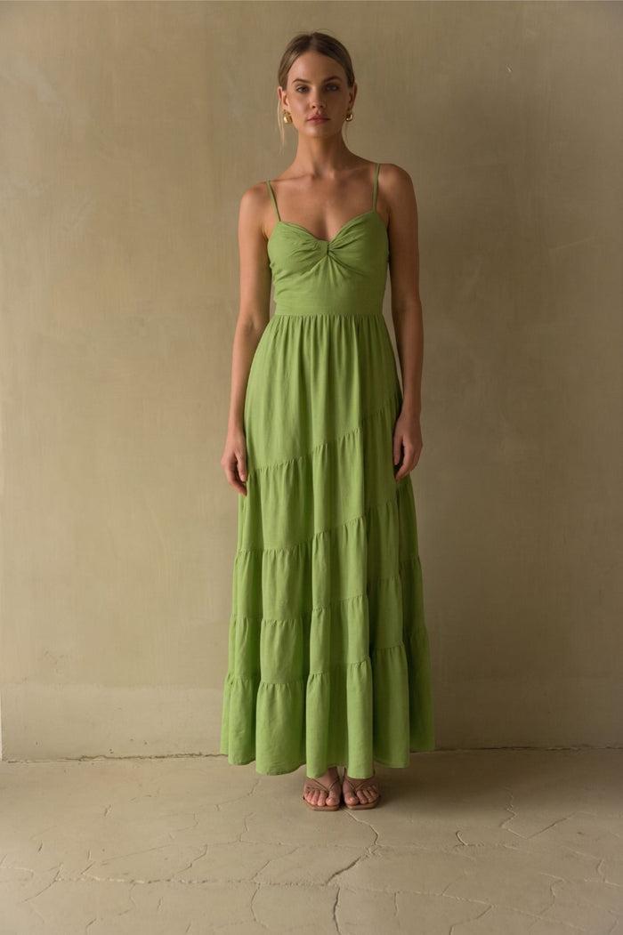 green-image | A woman stands barefoot against a textured beige wall wearing a sleeveless, tiered maxi dress in a light olive green color.
