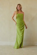 A woman modeling a strapless lime green maxi dress with draped detailing, standing against a textured beige plaster wall.